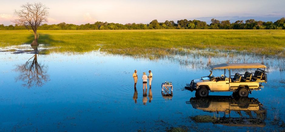 A safari vehicle is parked in a shallow, reflective floodplain where three people stand barefoot in the water, enjoying a sundowner with a small table set for drinks, surrounded by vast golden-green grasslands and a lone tree mirroring in the water.